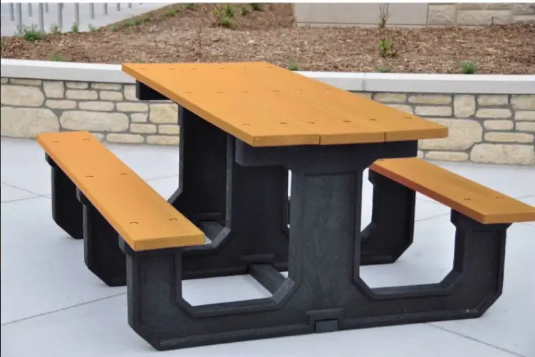 Rectangular recycled plastic picnic table with tan planks, attached benches, and a heavy-duty black frame in an outdoor courtyard.
